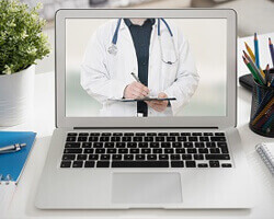A laptop computer on a white desk, with a doctor's torso in a labcoat on the display screen.
