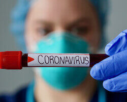 Close-up of a vial of blood labeled with "coronavirus," being held by a healthcare professional wearing protective gloves, mask, and headwear. 