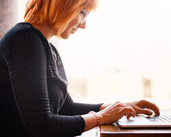 A red-haired woman seen from the side, sitting at a table and typing on a laptop computer. 