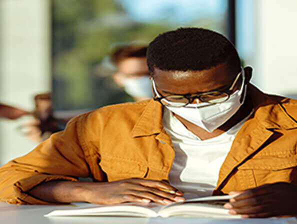 A young student with a PPE mask on reads a book at his school desk.
