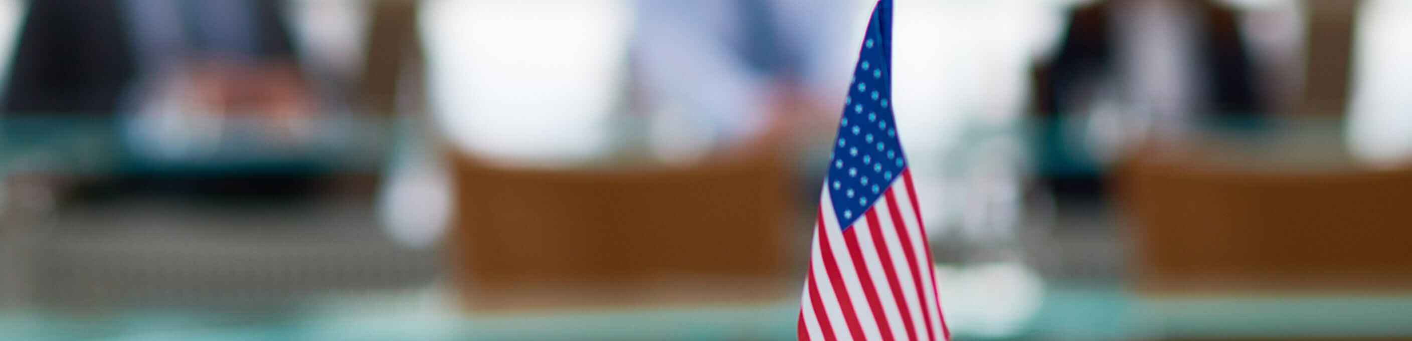 An American flag on a desk with meeting attendees blurred in the background. 