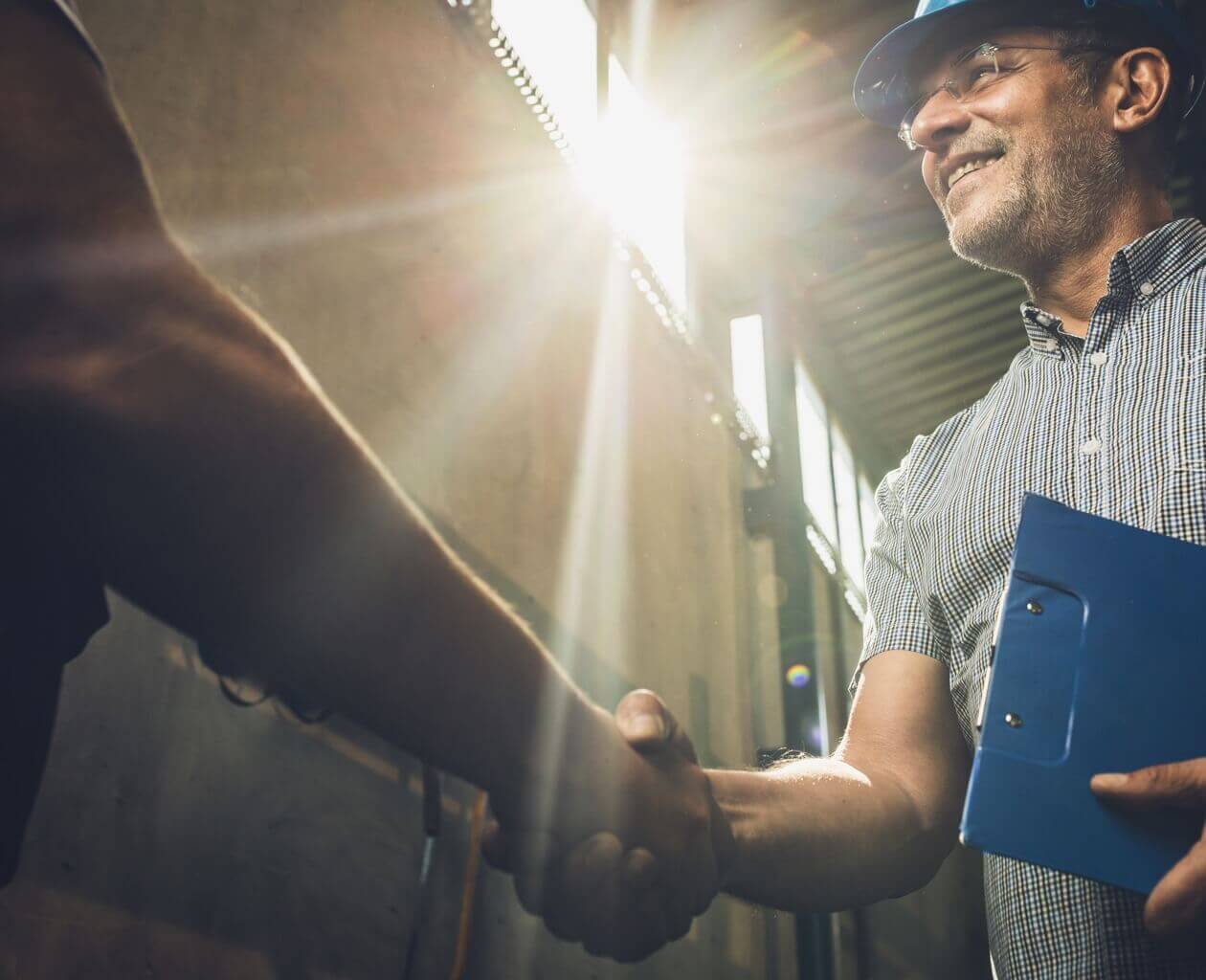 An engineer with a clipboard and hard hat shaking hands with someone in the sunshine. 