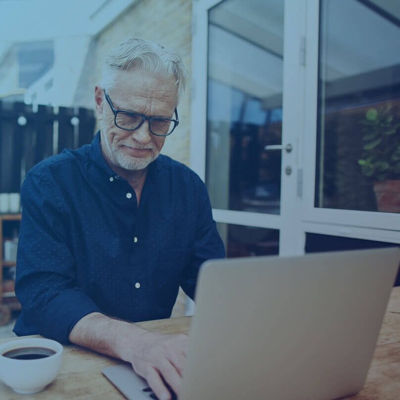An older Caucasian man in glasses with a cup of coffee, using his laptop and smiling. 