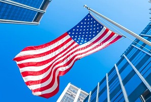 Upward view of an American flag flying with a bright blue sky and modern office buildings. 