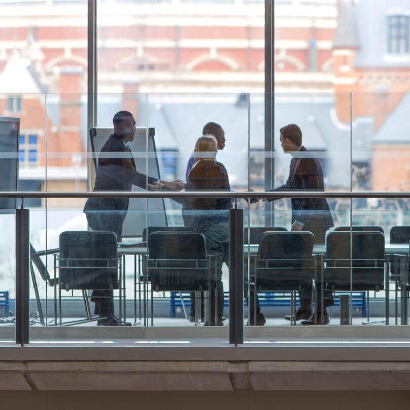 A small group of professionals seen from a distance through office windows, shaking hands and standing by a meeting table. 