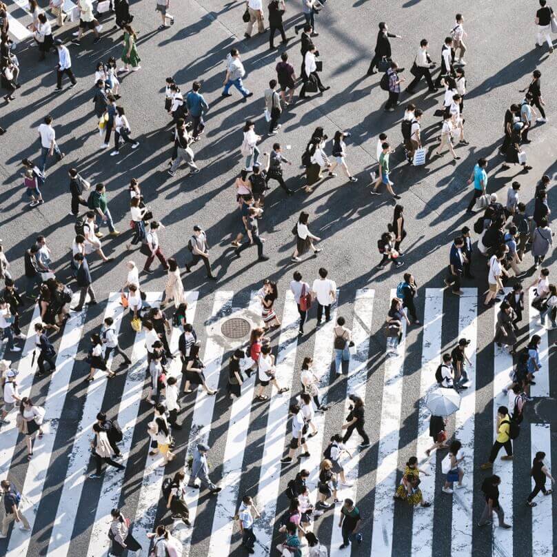 An aerial view of a large group of diverse people walking on a city street and in a crosswalk. 
