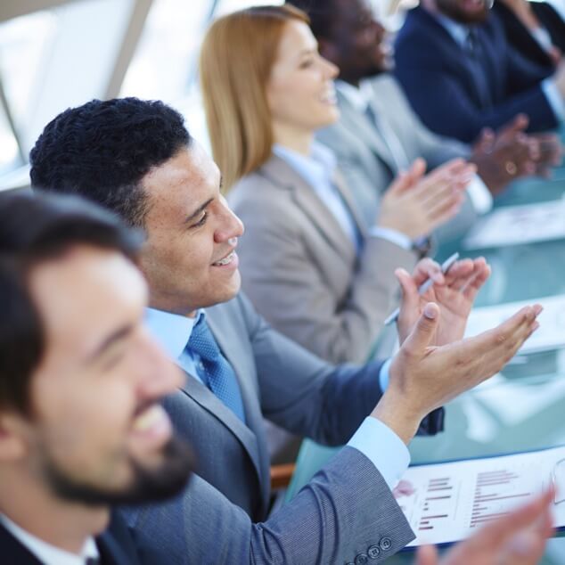 A row of four businesspeople sitting at a conference table clapping. 