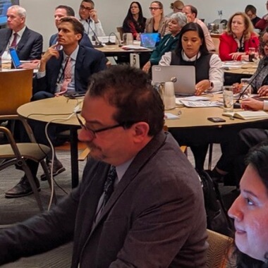 A seated businessman in a meeting room with a large group of other meeting attendees seated at tables in the background. 